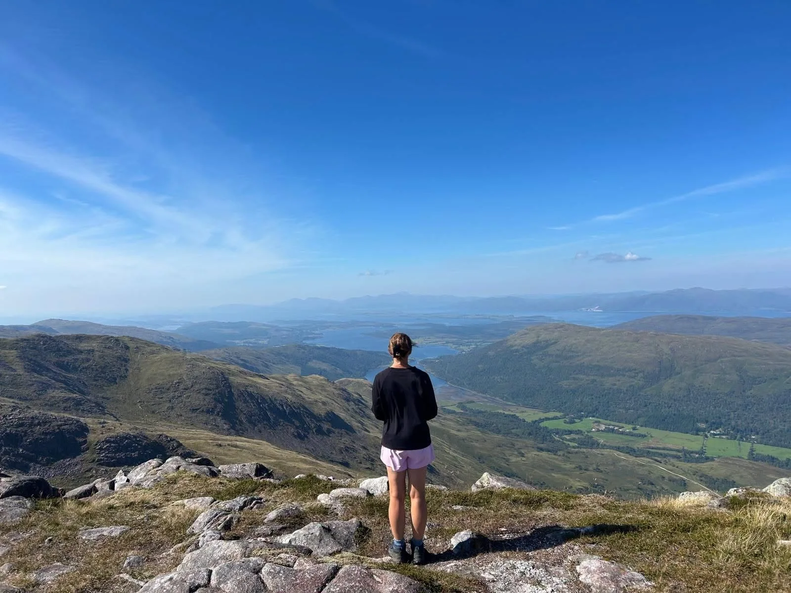 A hiker on a rocky summit looking out across mountains and lochs beneath a blue sky.
