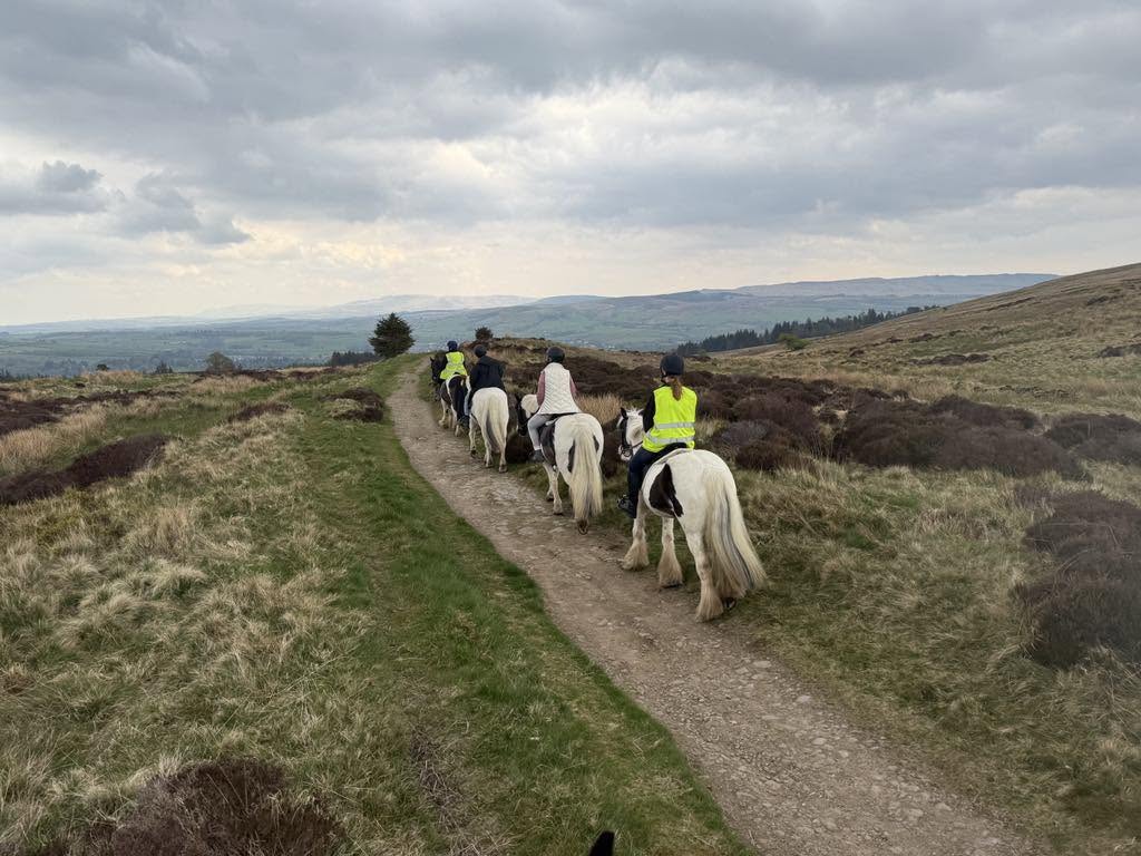 Riders on ponies following a trail across open moorland, with hills in the distance.
