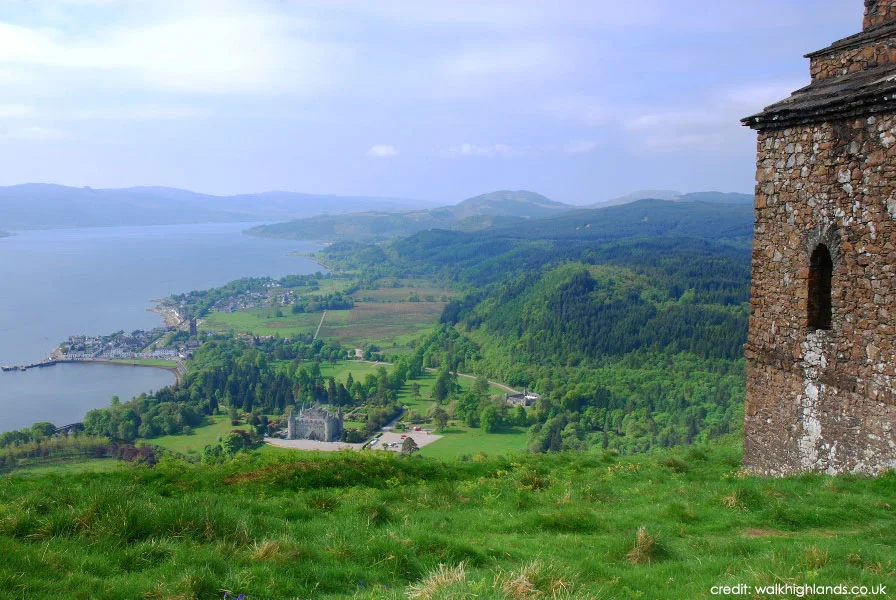 View of Inveraray Castle, a loch and wooded hills from a grassy summit beside a stone tower.