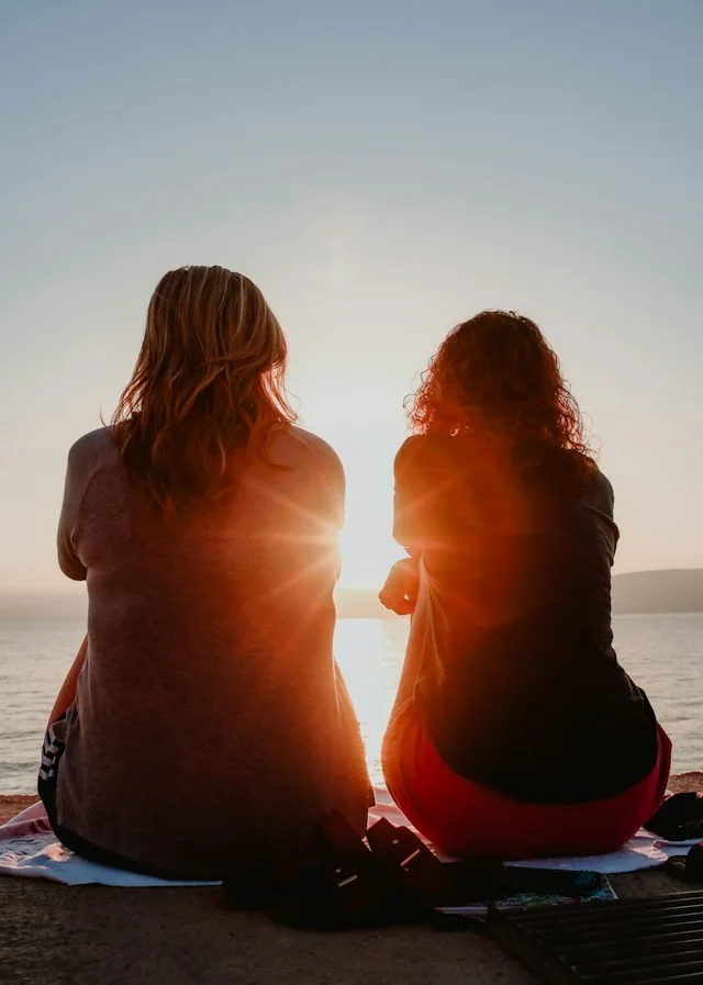 Two women sitting together beside the water at sunset.