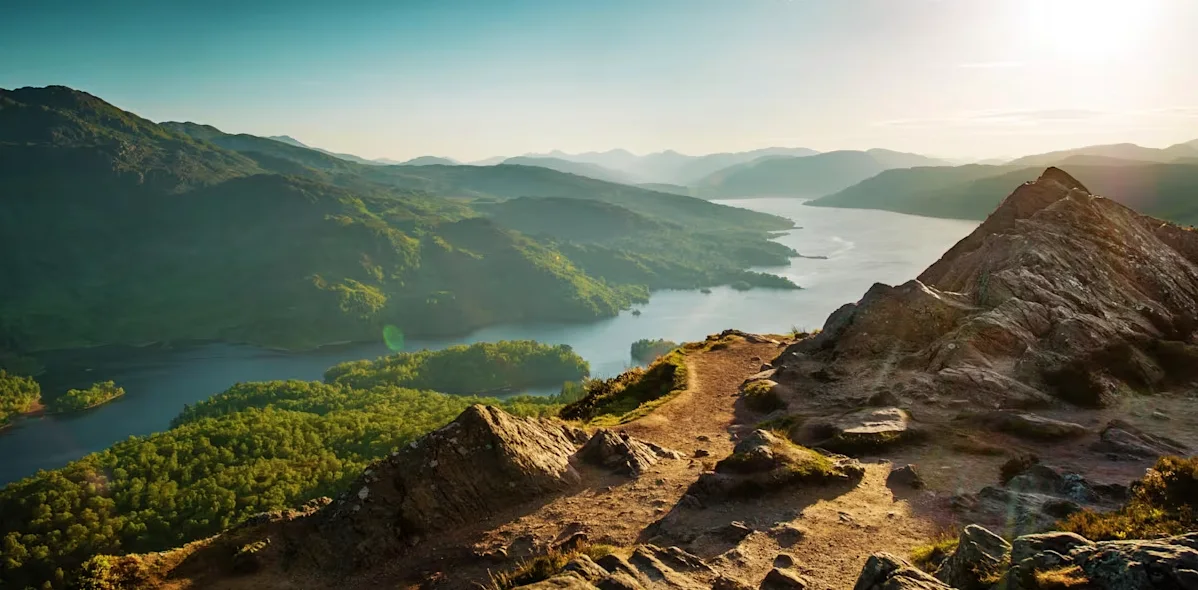 Sunlit rocky ridge overlooking a loch, wooded shores and distant hills.