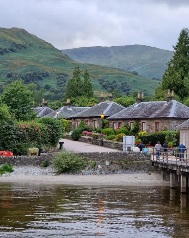 Stone cottages and gardens beside the water in Luss, with green hills behind.
