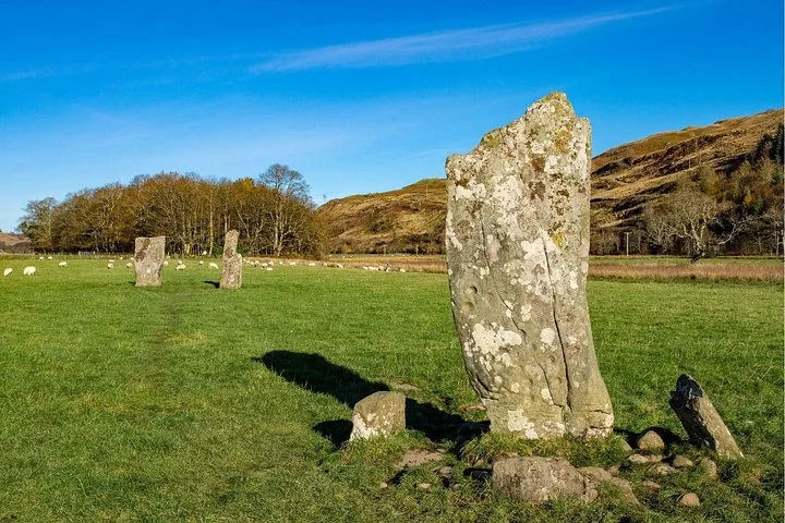 Lichen-covered standing stones in a grassy field, with trees and hills beneath a blue sky.